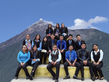 About 20 nicely dressed teenage kids sitting on a stair pyramid posing for a photo with a volcano in the background